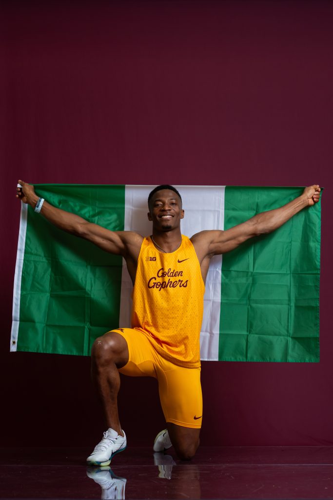 An athlete in a yellow "Golden Gophers" uniform kneels while holding up a Nigerian flag against a maroon background.